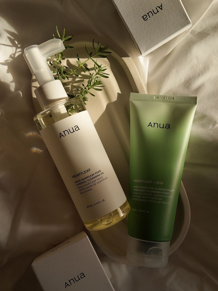 A flatlay composition featuring Anua skincare products, including a cleansing oil in a bottle and a green tube of pore cleansing foam, surrounded by soft lighting and a decorative plate with a sprig of greenery.